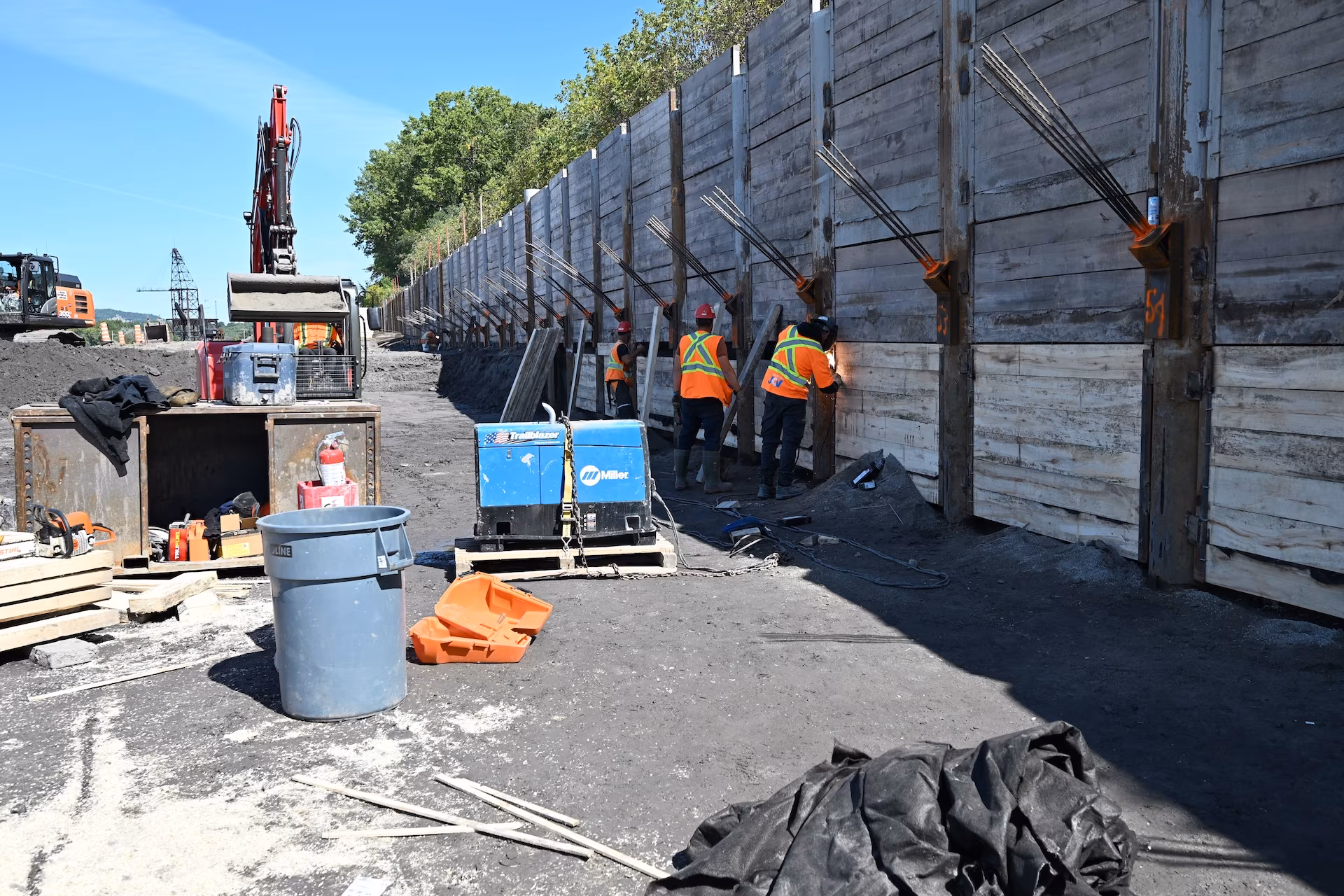 Construction workers in orange safety vests working on a reinforced retaining wall with heavy machinery and equipment nearby.