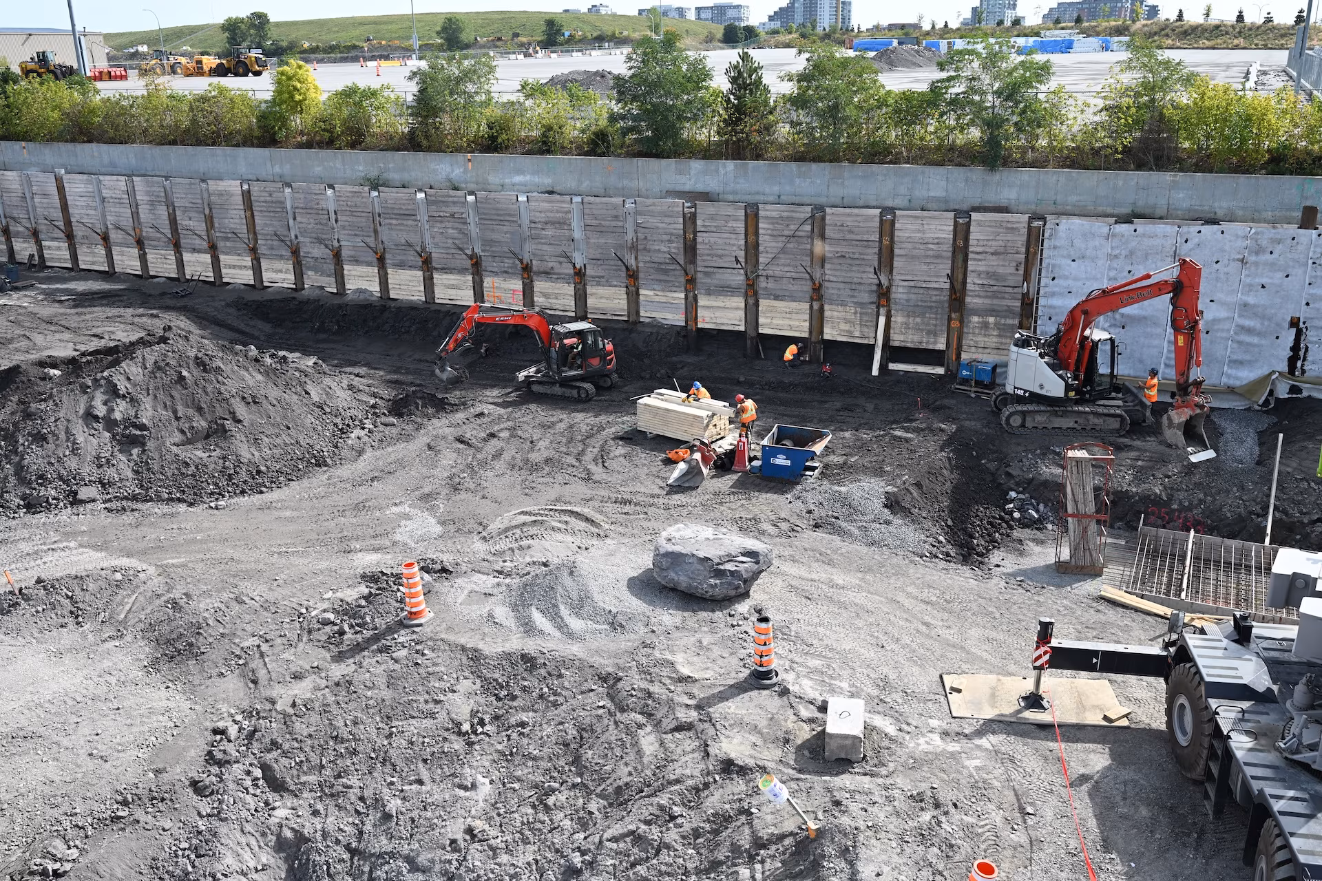 Construction site with workers in orange vests operating excavators and handling materials by a wooden retaining wall.