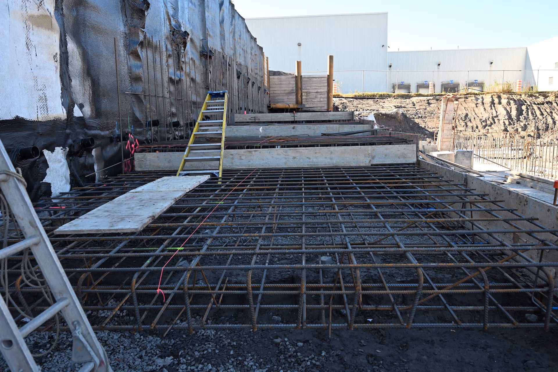 Construction site showing a steel rebar grid laid out for concrete foundation with ladders and wooden boards.
