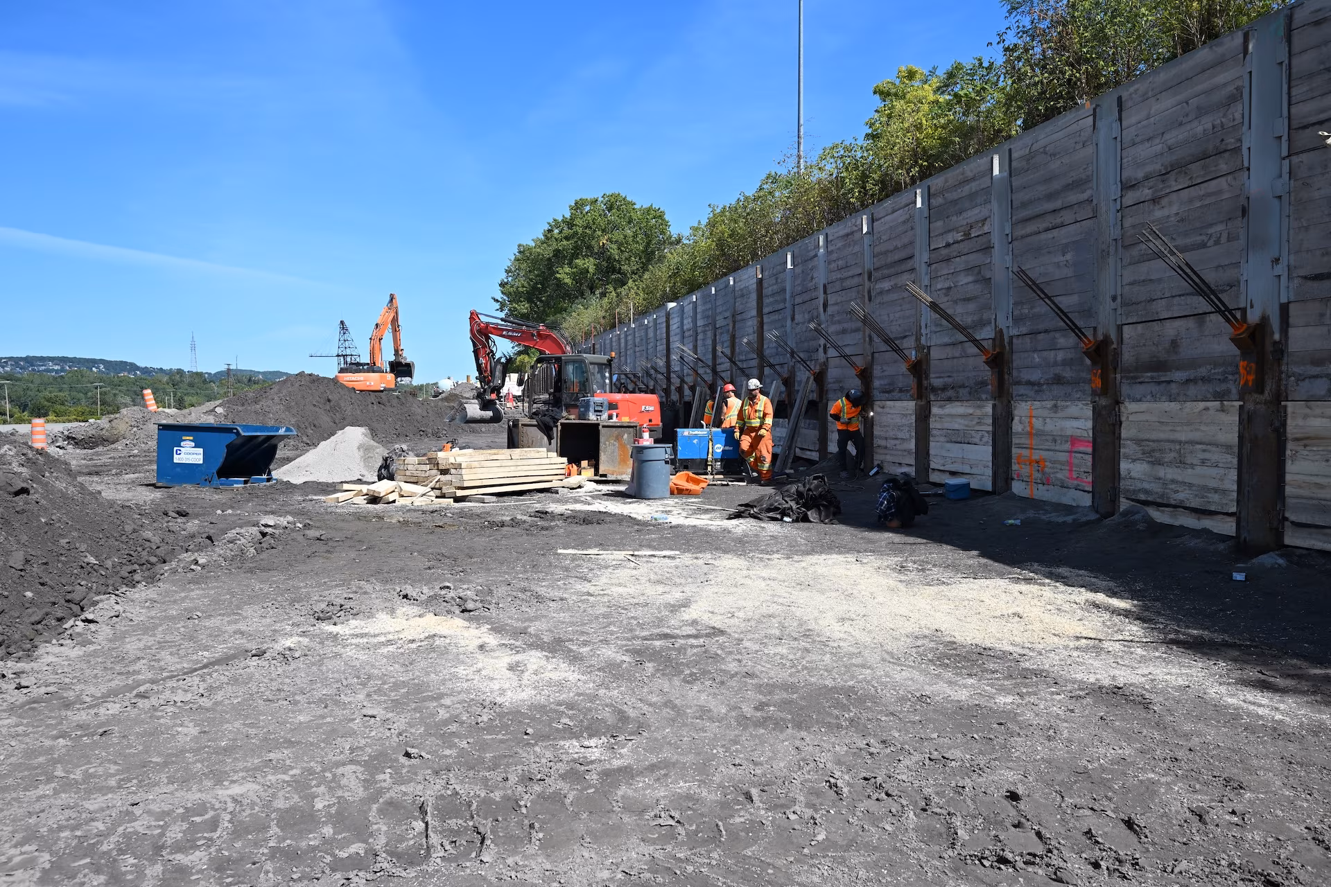 Construction site with workers in safety gear, machinery, piles of dirt, and a tall retaining wall with metal supports.